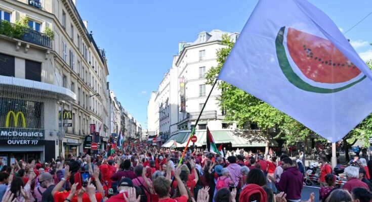 Egy fehér zászlót, amelyen egy nagy szelet görögdinnye van, a július 8 -i Párizsban zajló Gázai rendezvény piros vonalán izgatott. A legtöbb tüntető piros inget visel, és egy utcán sétálnak, amely több állammal bélelt épületekkel.